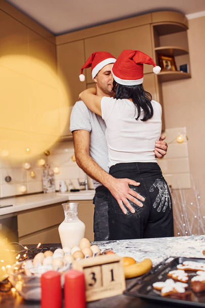 Couple with santa hats standing on the kitchen kissing and embracing each other. - Foto, Imagem