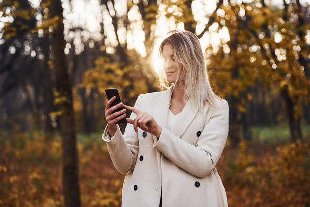 Portrait of young brunette with phone in hand that is in autumn forest at daytime. - Foto, Imagen
