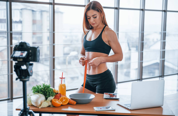 Feminino vlogger com corpo esportivo em pé dentro de casa com fita métrica perto da mesa com alimentos saudáveis. - Foto, Imagem