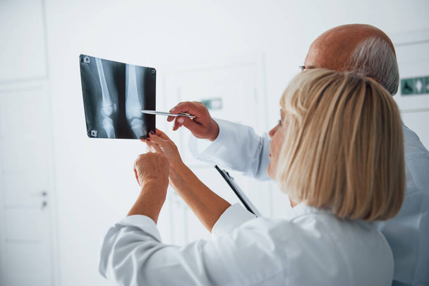 Senior man and woman doctors in white uniform examines x-ray of human legs. - Zdjęcie, obraz