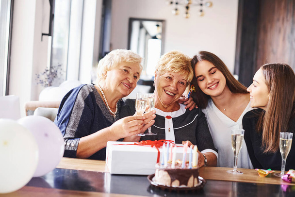 Okulary z alkoholem w rękach i tort na stole. Starsza kobieta z rodziną i przyjaciółmi świętującymi urodziny w domu. - Zdjęcie, obraz