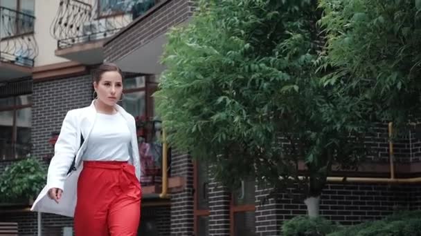 mulher morena confiante em calças vermelhas e blusa branca e casaco andando na rua. câmara lenta - Filmagem, Vídeo