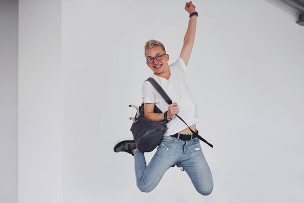Happy hipster guy in casual clothes and with backpack jumping indoors against white wall. - Valokuva, kuva