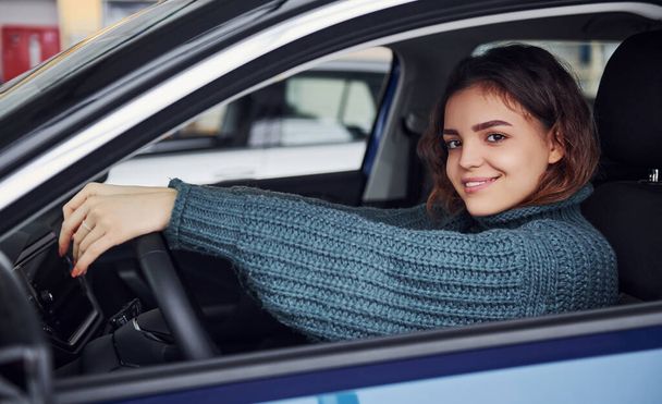 Positive young girl in casual clothes sitting inside of modern car and smiling. - Photo, image