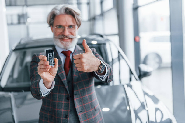 Senior businessman in suit and tie with gray hair and beard standing indoors near car with keys in hand. - Foto, Imagen