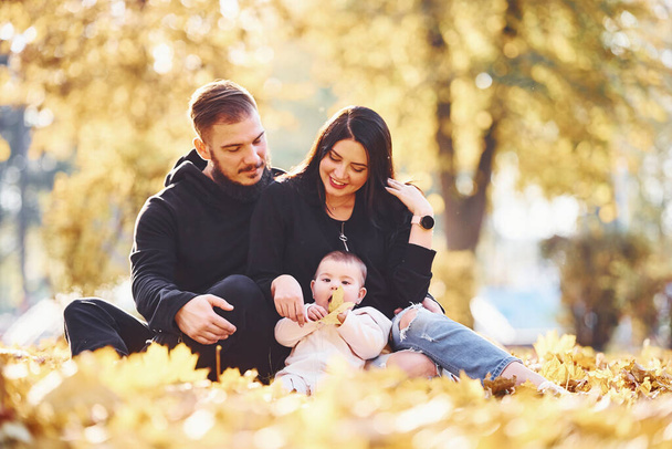 Cheerful family sits on the ground and having fun together with their child in beautiful autumn park. - Фото, зображення
