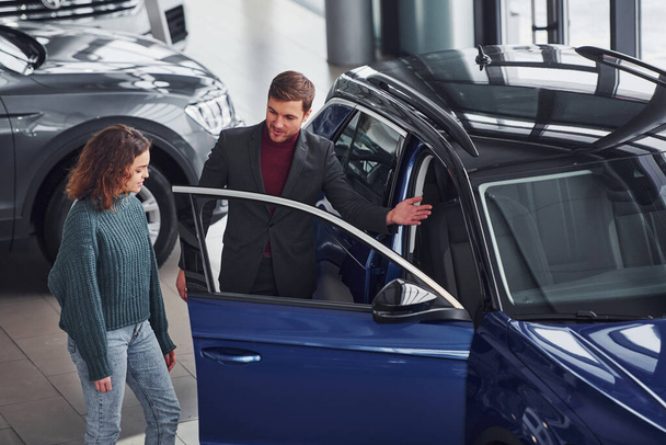 Professional salesman assisting young girl by choosing new modern automobile indoors. - 写真・画像