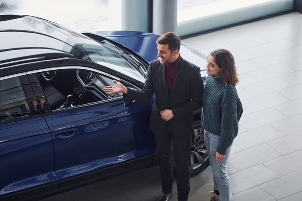 Professional salesman assisting young girl by choosing new modern automobile indoors. - Fotó, kép