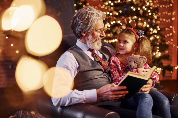 Cheerful fashioned senior man with grey hair and beard sitting with little girl in decorated christmas room reading book. - Foto, immagini