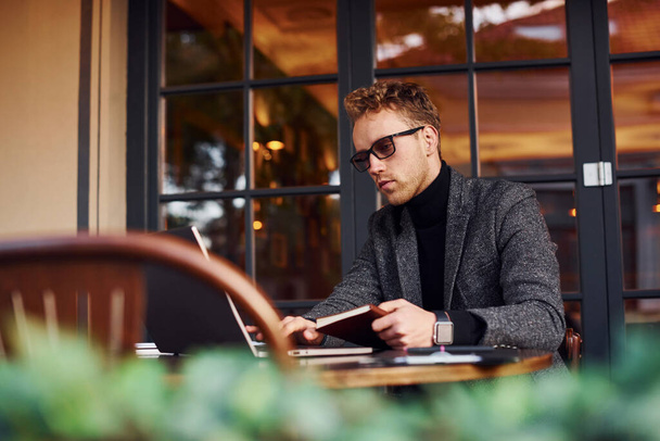 Elegant young guy in formal wear sits in cafe with his laptop and reads the book. - Foto, Imagem