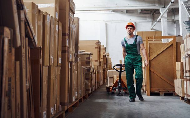 Young male worker in uniform is in the warehouse with pallet truck. - 写真・画像