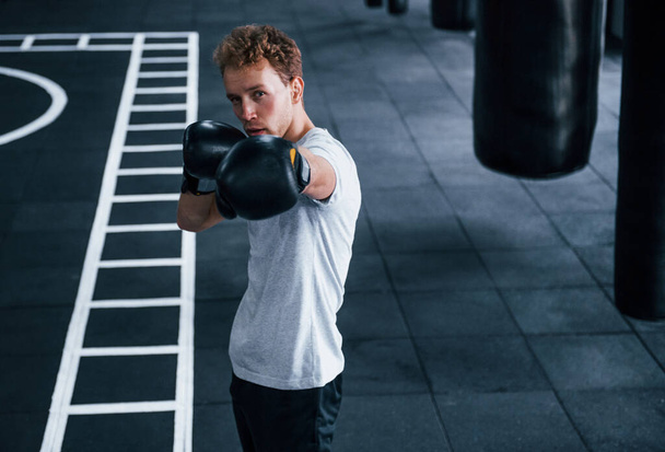 Young man in white shirt and boxing protective gloves doing exercises in gym. - 写真・画像