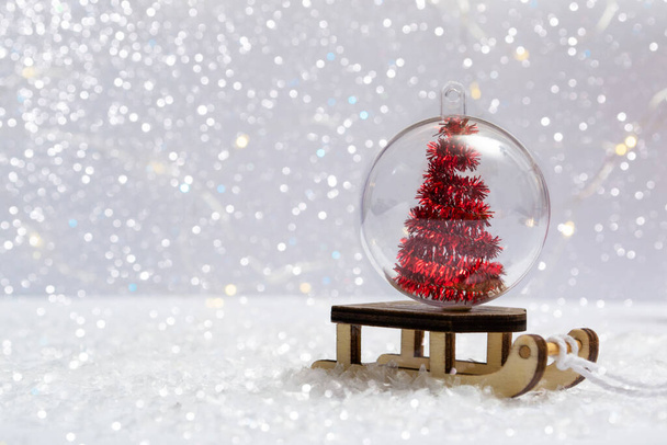 Sulla neve - una slitta con una palla di Natale dentro un albero di Natale su uno sfondo di luci di bokeh primo piano. Foto verticale - Foto, immagini