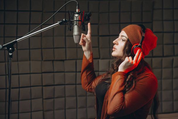 Young woman in headphones emotionally singing in microphone in the sound recording studio. Star, talent, preparing show, studio recording. Soundproof room for professional recording vocal - Zdjęcie, obraz