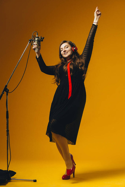 The positive amusing brunette female singer having fun sings song. Young female caucasian pop artist in earphones smiling while performing joyful song in microphone on the yellow background - Photo, Image