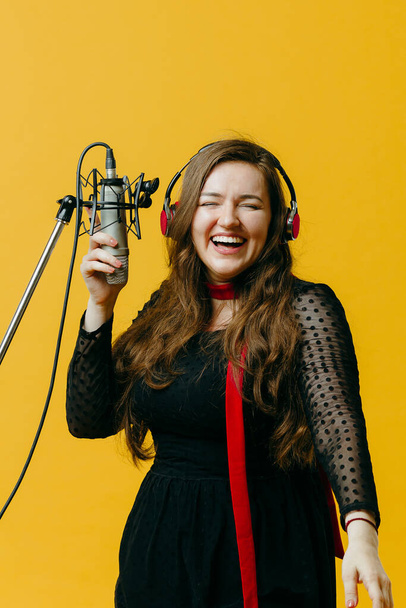 The positive amusing brunette female singer having fun sings song. Closeup of young female caucasian pop artist in earphones smiling while performing joyful song in microphone on the yellow background - Foto, afbeelding