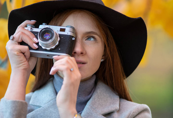 Portrait of redhead girl with hat taking photographs with old analog camera in park in autumn time - Photo, Image