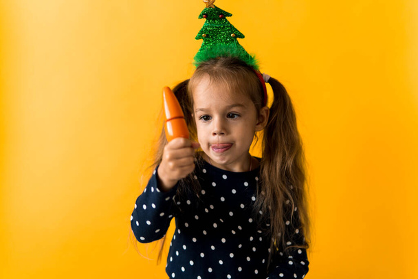 Portrait positive cheerful smiling happy little schoolgirl girl Christmas tree decoration polka dot dress biting eat orange carrots on orange background. New year, holiday, celebration, winter concept - Фото, зображення
