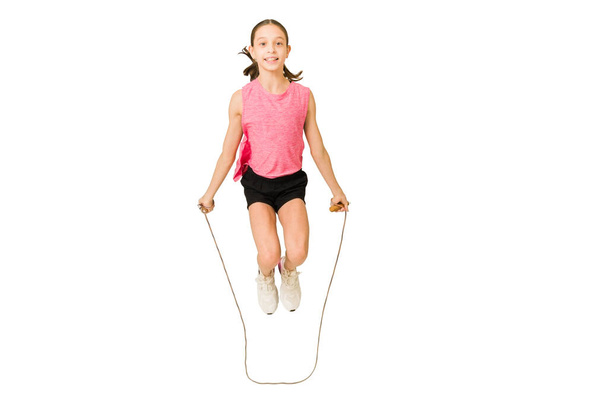 Alegre niño pequeño en ropa deportiva sonriendo y disfrutando saltando la cuerda sobre un fondo blanco - Foto, imagen