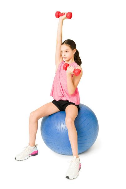 Attivo giovane ragazza sollevamento manubri pesi sopra la testa durante l'utilizzo di una palla fitness - Foto, immagini