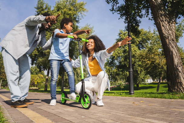 Beautiful happy african american family bonding at the park - Black family having fun outdoors, parents teaching son to ride on scooter - Photo, Image