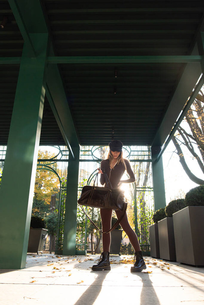 Модная молодая брюнетка с длинными ногами носить Твердый длинный рукав Bodycon One Piece Jumpsuits позировать с кожаной коричневой дорожной сумкой - Фото, изображение