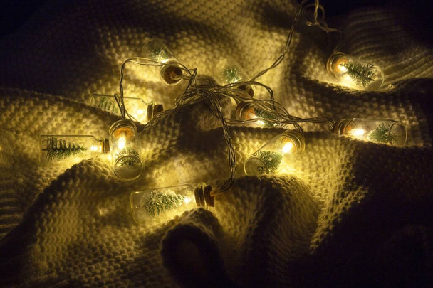 クリスマスツリーと人工雪の入ったガラスボトルで作られた美しい手作りの工芸品クリスマスツリーガーランドは、白いニットセーターに横たわっています。クリスマスの週末の快適さと居心地のよさ. - 写真・画像