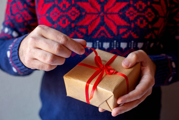 A man in a sweater with a New Year's pattern holds a Christmas present on a grey background. Gift concept for Christmas and New Year. - Photo, Image