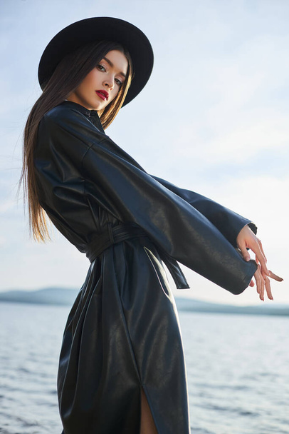 Fashion woman in black round hat and leather raincoat near lake pond. Portrait girl at sunset. Beautiful makeup and bright red lipstick on lips - Фото, зображення