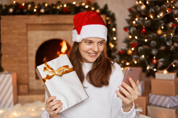 Indoor shot of happy positive woman wearing white sweater and santa claus hat, posing near fireplace and xmas tree, having video call, showing her new year present to gadget camera. - Fotografie, Obrázek