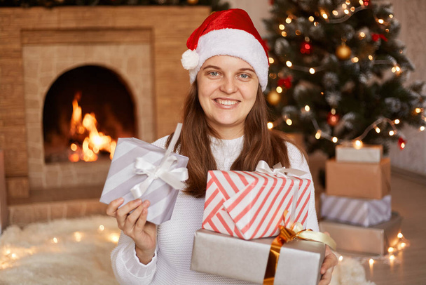 Smiling satisfied woman wearing white sweater and santa claus hat, holding gift boxes, congratulating with winter holidays, being in festive room with fireplace and Christmas tree. - Fotó, kép
