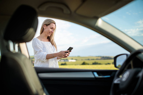 Pretty middle aged woman at the steering wheel of her car, having a break on a long trip, making a phone call - Photo, image