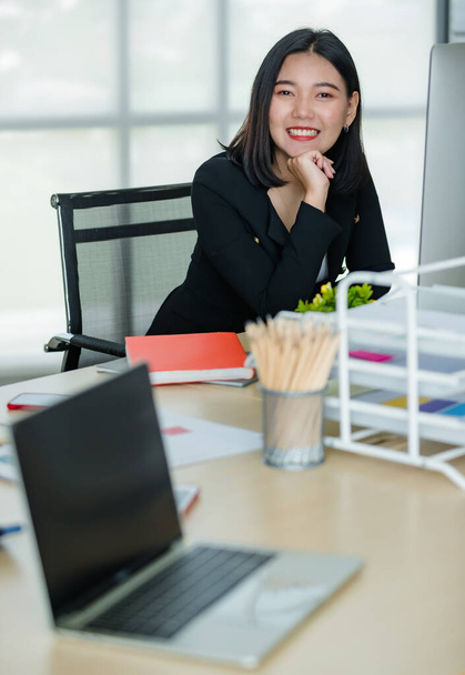 Giovane donna asiatica attraente in abito da lavoro nero seduto in ufficio moderno a lavorare e sorridere con il computer portatile sul tavolo. Concetto per uno stile di vita moderno. - Foto, immagini