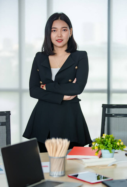 Mladá atraktivní asijská žena v černém obleku stojící ruku zkříženou s důvěrou v moderní vypadající kancelář. Koncept pro moderní kancelářský životní styl. - Fotografie, Obrázek