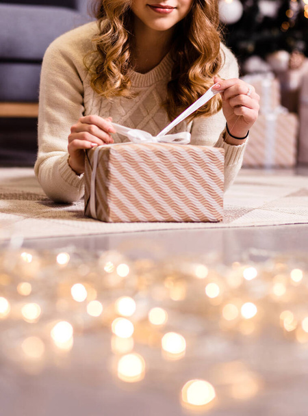 Une fille en pull blanc se couche sur le sol et ouvre un cadeau. Ambiance Nouvel An, bokeh doré au premier plan. Photographie - Photo, image