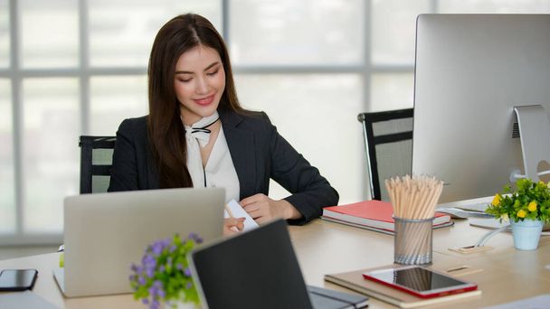Młoda atrakcyjna Azjatka w czarnym biznesie siedzi za stołem z komputerem i laptopem pracując w nowoczesnym biurze z rozmytymi oknami w tle. Koncepcja nowoczesnego stylu życia biurowego. - Zdjęcie, obraz