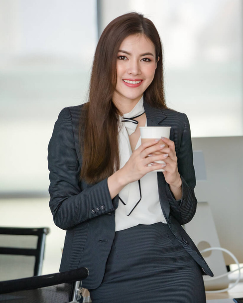 Siyahlar içinde genç çekici bir Asyalı kadın elinde bir fincan kahveyle modern görünümlü ofiste gülümsüyor ve arka planda bulanık pencereler var. Modern ofis yaşam tarzı kavramı. - Fotoğraf, Görsel