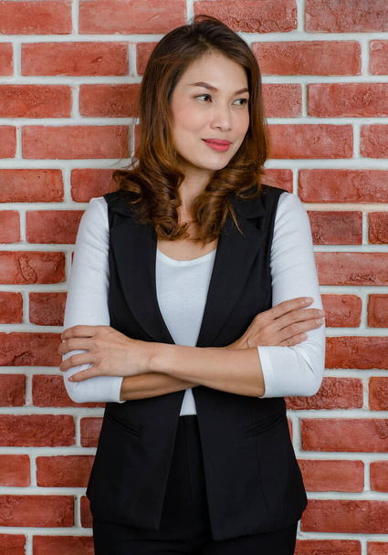 Orta yaşlı çekici Asyalı kadın portresi siyah yelek ve beyaz tişörtle turuncu tuğla arka planda poz veriyor ve gülümsüyor.. - Fotoğraf, Görsel