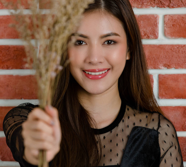 Close-up de jovem bonita atraente modelo feminino asiático superior torso sorrindo segurando ramo da flor contra fundo de parede de tijolo laranja. - Foto, Imagem