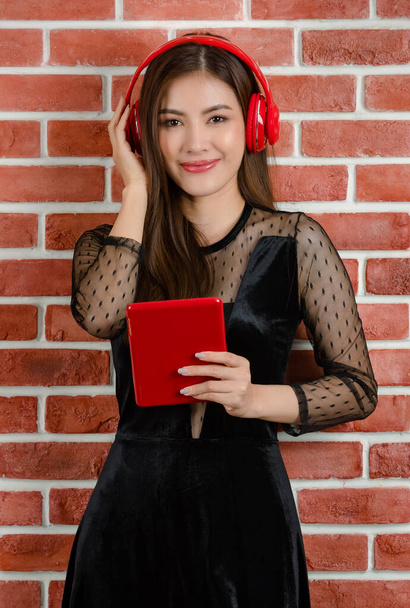 Portrait of young beautiful attractive Asian female model in black revealing dress wearing red headphone listening to music and holding tablet against orange brick wall background. - Photo, Image