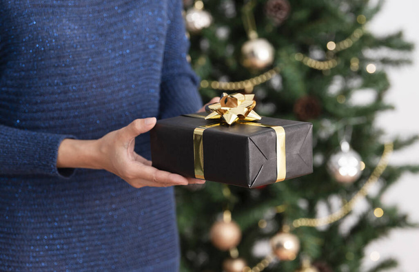 Woman in sweater holding gift box near christmas tree background. - Фото, изображение