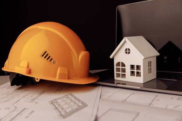 Желтый шлем, дом и ноутбук со строительными планами на столе - Фото, изображение