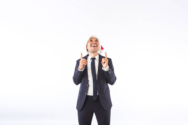 Businessman dressed in suit, tie and Christmas Santa hat, smiling, pointing up with his hands, on white background. Christmas celebration concept. - Photo, Image