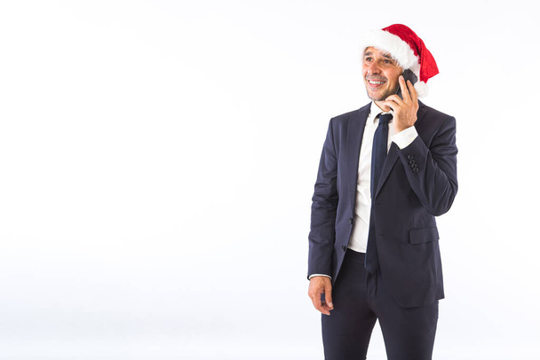 Empresario vestido de traje, corbata y sombrero de Navidad de Santa Claus, hablando por teléfono, sobre fondo blanco. Concepto de celebración de Navidad. - Foto, Imagen