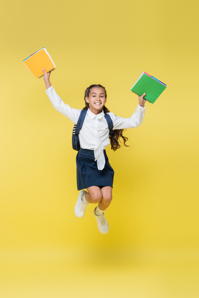 Happy schoolkid with backpack and notebooks jumping on yellow - 写真・画像