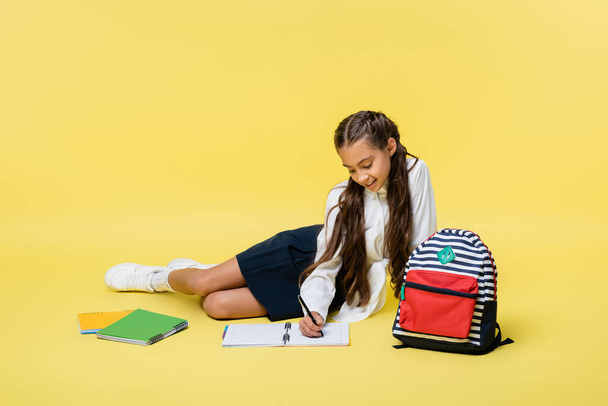 Smiling kid writing on notebook near backpack on yellow background  - 写真・画像