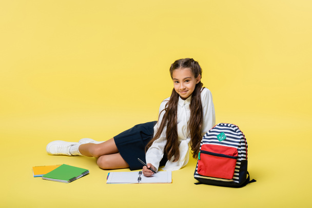 Schoolgirl writing on notebook and smiling at camera near backpack on yellow background  - Foto, Bild