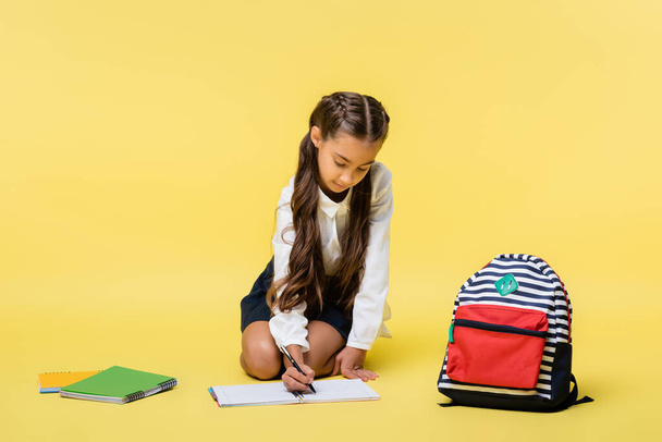 Schoolgirl writing on notebook near backpack on yellow background  - Fotó, kép