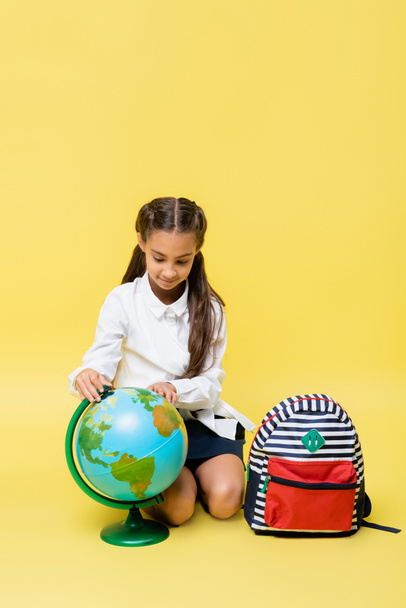 Schoolchild looking at globe near backpack on yellow background  - Фото, изображение
