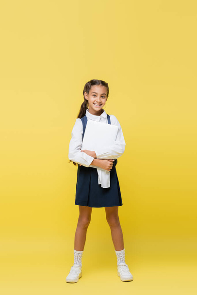 Full length of smiling schoolgirl with backpack holding laptop on yellow background - Photo, Image